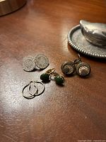 Top-down view of four pairs of earrings including silver handmade filigree, gold drop earrings with green stones, vintage button clip-ons, and thin gold hoops.