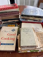 Stack of various vintage and newer cookbooks and handwritten recipe books displayed on a table, showing worn covers and aged paper.
