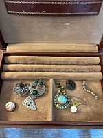 The jewelry box with various costume brooches and a clip earring placed inside, showing the collection style and box interior.