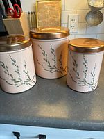 Front view of three pink metal nesting canisters decorated with pussy willow branches, placed side by side on a countertop with gold lids on top.