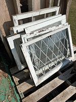 Six vintage wooden window frames leaning against wooden pallets outside, showing various glass styles and chipping white paint on the frames.