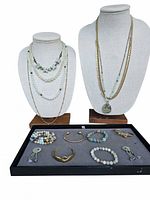 Photo showing two bust stands displaying multiple gemstone and beaded necklaces in seafoam green and gold tones, with a tray below holding bracelets and small charms.