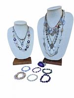 Two jewelry busts displaying layered necklaces in tones of purple, grey, and blush pink, with bracelets laid flat below in shades of purple and pearl.