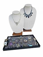 Two bust display mannequins holding two distinct necklaces; one with dark beaded chain and multiple leaf-shaped mother of pearl pendants, the other simpler silver-tone chain with small mother of pearl pendant. Lower tray displays a variety of pendants, charms and bracelets made from mother of pearl and metal.