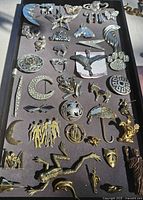 Silver and gold toned vintage pins and brooches arranged on tray surface, displaying diverse shapes including animals, celestial, abstract and symbolic motifs
