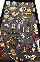 Top view of vintage pins and brooches arranged in a tray showing a variety of shapes, sizes and materials such as gold-tone, silver-tone, faux pearls, and red enamel accents.