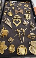 Top view of assortment of vintage gold-tone pins and brooches arranged on black tray showing various animal, floral, and heart designs.