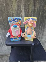 Photo of two vintage Ren & Stimpy plush toy dolls still attached in original packaging showing full front view on a black stand against a concrete background.