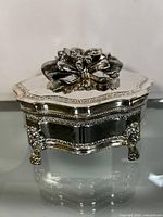 Close front view of silver-tone jewelry box showing floral bow on lid and ornate legs