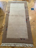 Full view of the rectangular wool rug showing the beige center panel, brown border with geometric accents, and fringed edges.
