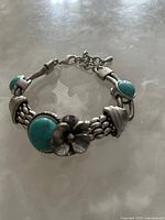 Photo showing silver-tone bracelet with woven chain links, turquoise stones, and flower accent lying flat on a surface.