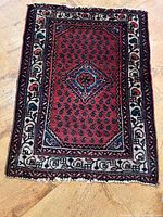 Top-down view of the Persian wool carpet showing the red field with blue diamond medallion and ivory floral borders.