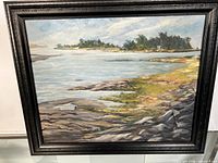 Framed oil painting showing a wide view of rocky shoreline, water, and distant trees and building.