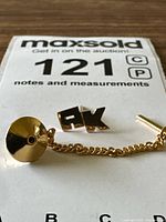 Close-up view of both front and chain backing disc details of the 14k gold tie pin with AK initials.