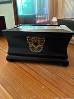 Closed view of black antique style jewelry box with clear glass lid and decorative gold-tone metal on front, sitting on wooden surface
