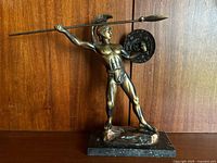 Front view of the bronze Roman warrior statue with spear and shield on marble base against wooden background.