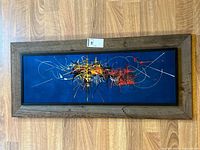 Full view of framed abstract art print on wooden floor showing natural wood grain frame and blue background with colorful abstract lines and shapes.