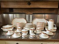 Full view of the dinnerware set including mugs, plates, bowls, serving platters, covered dish, and tall pitcher with lid arranged on a counter.