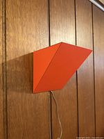 Single red metal sconce mounted on wood-paneled wall, showing angular V shape and hanging wire.