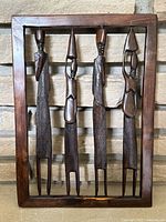 Full view of four hand-carved ebony wooden tribal figures mounted in a rectangular wooden frame against a stone background.
