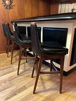 Side angled view of four black vinyl upholstered bar stools with wooden legs and metal foot bars lined up at a bar.
