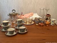 Wide view showing antique Adams Ironstone cups and saucers, clear stemmed glassware, various decorative pieces including ceramic and glass items on wooden table.
