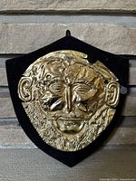 Front view of gold-tone metal mask depicting Agamemnon mounted on shield-shaped black velvet backing.