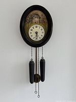 Full view of vintage wall clock with hand-painted scenic background, black oval frame, clock face, and hanging weights.