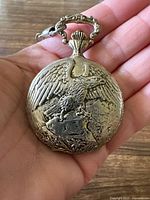 Front side of silver tone pocket watch with eagle in relief on the case, held in hand to show size and detail.