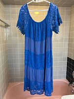 Full front view of blue tiered lace dress hanging on white hanger in tiled bathroom.