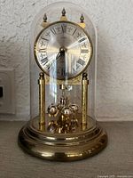 Clock inside clear acrylic dome showing brass finish, dial with Roman numerals, decorative finials and spherical pendulum.