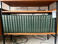 Full set of 23 dark green hardcover encyclopedia volumes with gold embossing, showing spines aligned on a wooden shelf, volume 18 missing.