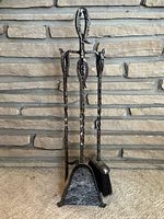 Full set of vintage pewter tone wrought iron fireplace tools including shovel, brush, poker, and stand in front of stone background
