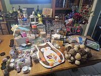 Wide view of assorted craft supplies arranged on a table including jars of pigments or glitters, paint bottles, storage containers, decorative pine cones, artificial flowers, buttons, and various craft papers and tools.
