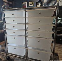 Front view of rolling metal craft table showing 6 white plastic drawers with metal knobs on each side in a silver metal frame.