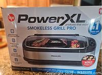 Box front showing PowerXL Smokeless Grill Pro branding, an image of the grill with food, and mentions bonus griddle plate included.