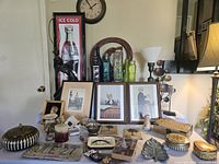 Wide view of collection of vintage household items including framed artwork, decorative glass bottles, several candle holders, small containers, platters, and a framed Coca-Cola sign leaning against the wall.