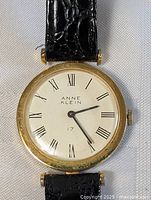 Close-up photo showing the white dial of the Anne Klein watch with black Roman numeral markers, gold-tone bezel, and black leather strap.