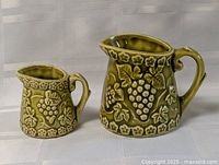 Both vintage ceramic measuring cups in olive green with grape and leaf embossed pattern shown side by side on white background.