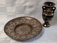 Photo showing hand hammered copper wall plate with floral and geometric etched designs next to Greek vase