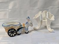 Photo shows two ceramic flower pot holders on a white cloth background. One is a donkey pulling a cart with black wheels, the other is a white calla lily shaped holder with three trumpet-like openings.