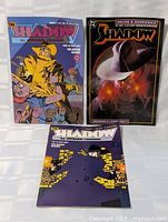 Three Shadow comic books arranged standing and lying flat on clear surface, showing their front covers clearly