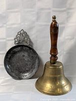 Photo showing brass school bell with a turned wooden handle next to a round pewter dish with ornate handle.