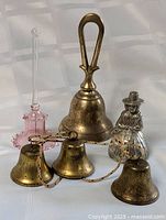 Photo showing a large solid brass bell with looped handle, five smaller brass bells connected by rope, and a clear glass figurine in the shape of a person.