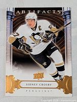 Front image of a Sidney Crosby hockey card from the Upper Deck Artifacts series showing player in white Penguins uniform with gold foil accents and logos.