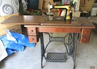 Full view of antique Singer treadle sewing machine on wooden table with drawers and black cast iron treadle base with 'SINGER' branding.