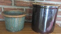Photo showing both planters side by side on a wooden surface against a brick background. The left is a blue-green textured seaweed planter with a rough brown rim, the right is a darker brown glossy antique crock with a beige rim.
