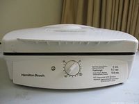 Front view of white Hamilton Beach contact grill showing timer dial with cooking times labeled for beef, hamburger, chicken breast, and hot dogs.