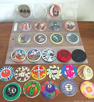 Photo of vintage Pogs and Slammers arranged in a plastic binder sleeve, showing various themes including Batman, cartoon characters, and colorful abstract designs.