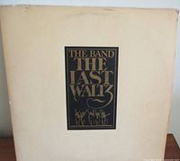 Front cover of The Band The Last Waltz vinyl LP album showing the title in gold and black lettering on a white background, slight discoloration visible.
